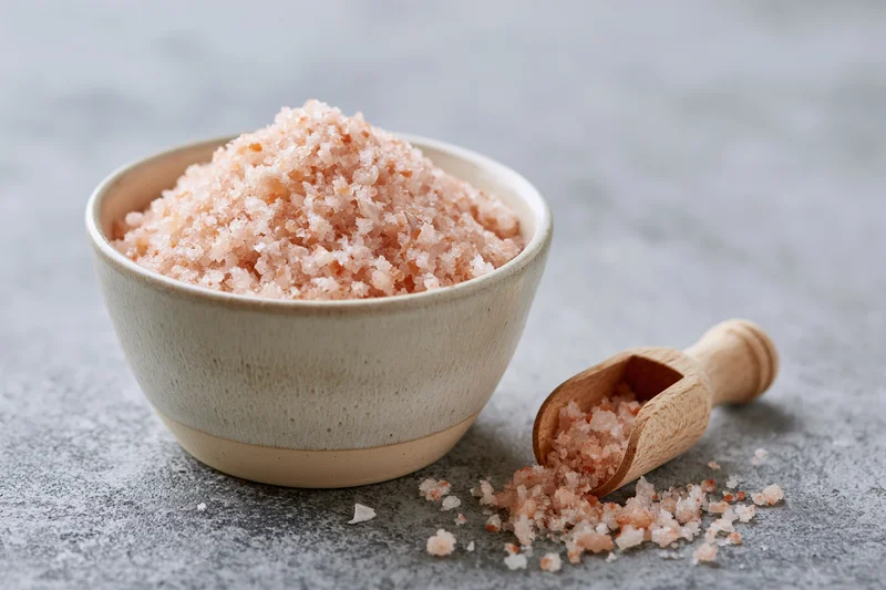 Ceramic bowl filled with pink himalayan salt beside a wooden scoop on a gray textured surface