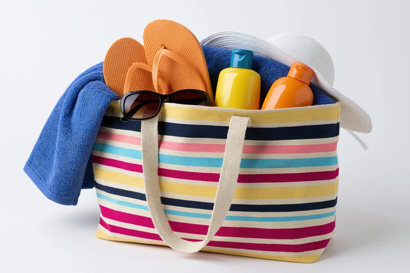 Colorful striped canvas beach bag filled with flip flops, sunglasses, towel, sunscreen bottles, and hat on white background