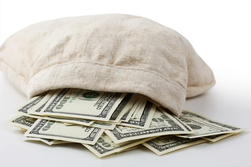 Cream canvas money bag tipped on its side spilling US hundred-dollar bills onto white surface
