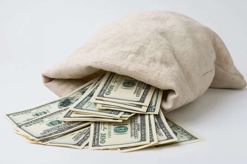Cream canvas bag tipped over spilling hundred-dollar bills on white surface
