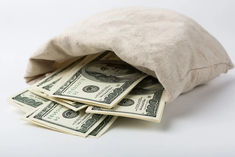Cream canvas money bag tipped on its side spilling hundred-dollar bills onto white surface