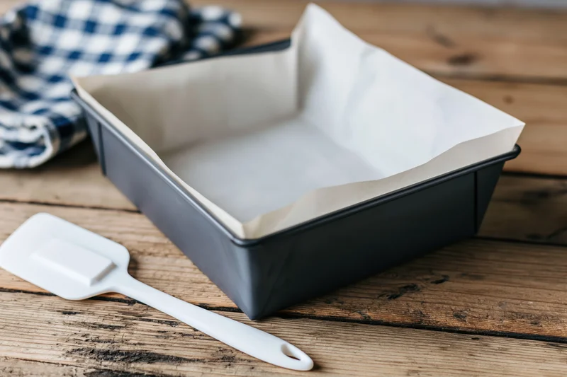 Dark gray metal loaf pan lined with white parchment paper on wooden table with white spatula and checkered cloth nearby