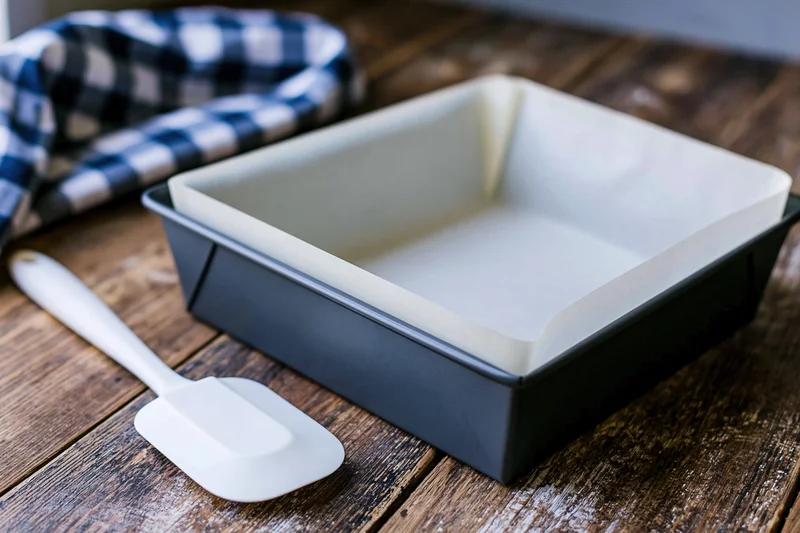 Dark grey metal loaf pan lined with white parchment paper on wooden table with white spatula and checkered cloth nearby