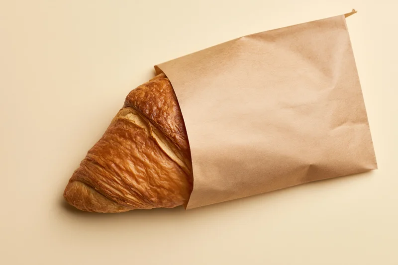 Golden croissant partially wrapped in kraft paper on beige surface