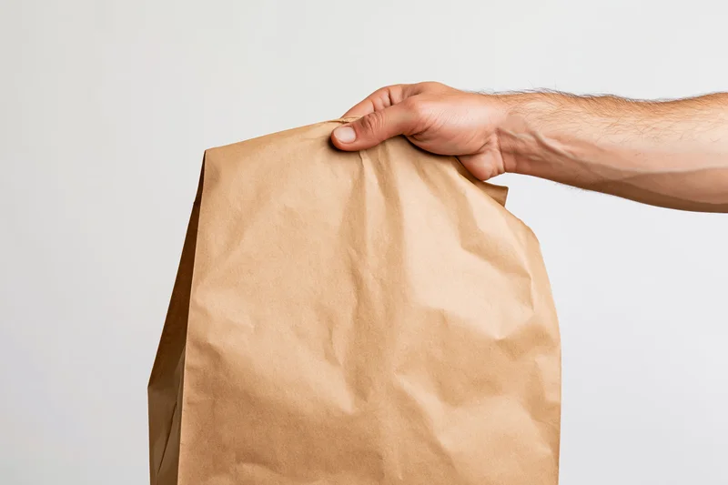 Hand gripping the top of a plain kraft paper takeout bag against a white background