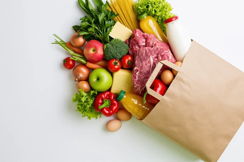 Kraft paper grocery bag tipped on its side spilling fresh produce, meat, dairy and pantry staples onto white surface