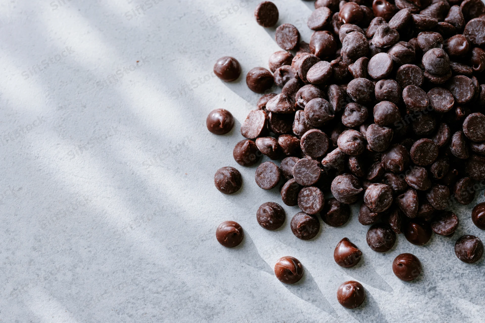 Chocolate chips scattered on a gray surface with natural light and shadows, copyspace on left