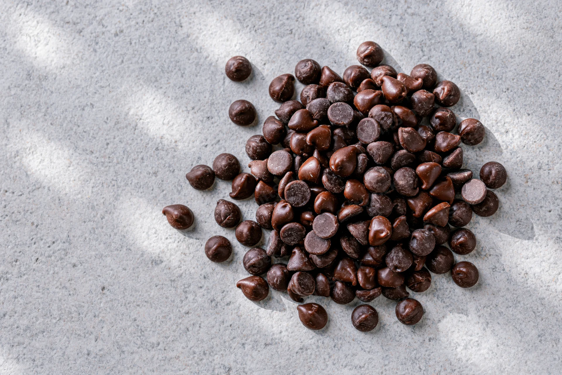 Pile of semi-sweet chocolate chips scattered on a gray textured surface with natural light and shadows