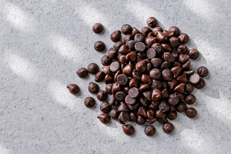 Pile of semi-sweet chocolate chips scattered on a gray textured surface with natural light and shadows