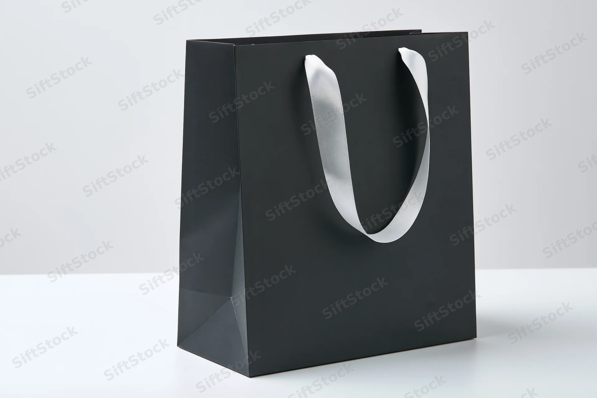 Matte black paper shopping bag with white ribbon handles on white surface
