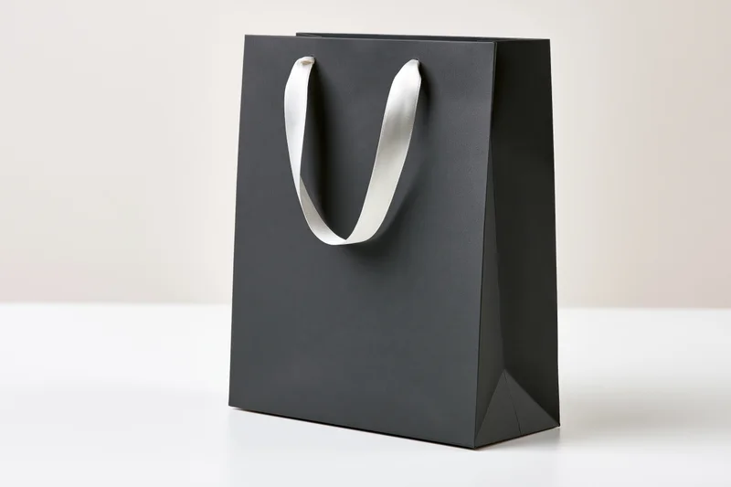 Matte black paper shopping bag with white ribbon handles standing on white surface