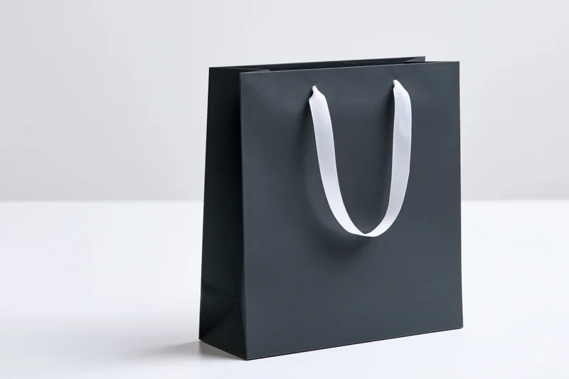 Matte black paper shopping bag with white ribbon handles on white surface