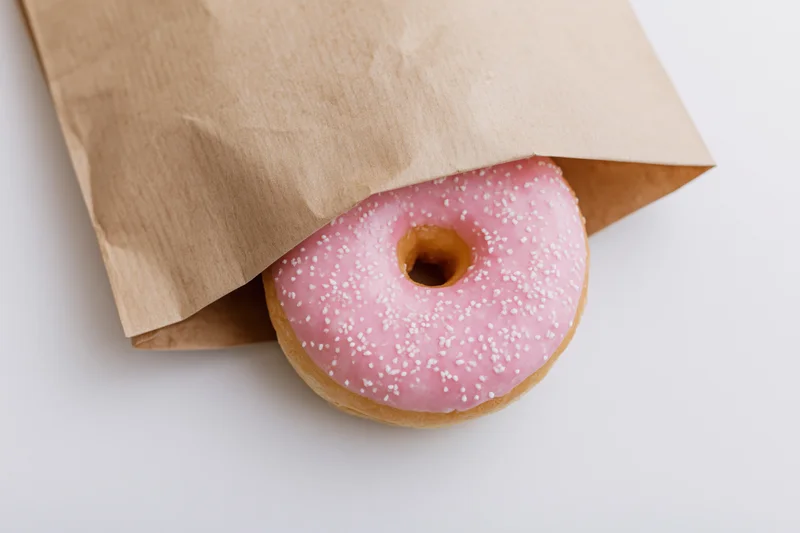 Pink glazed donut with white sprinkles peeking from a brown paper bag on white surface