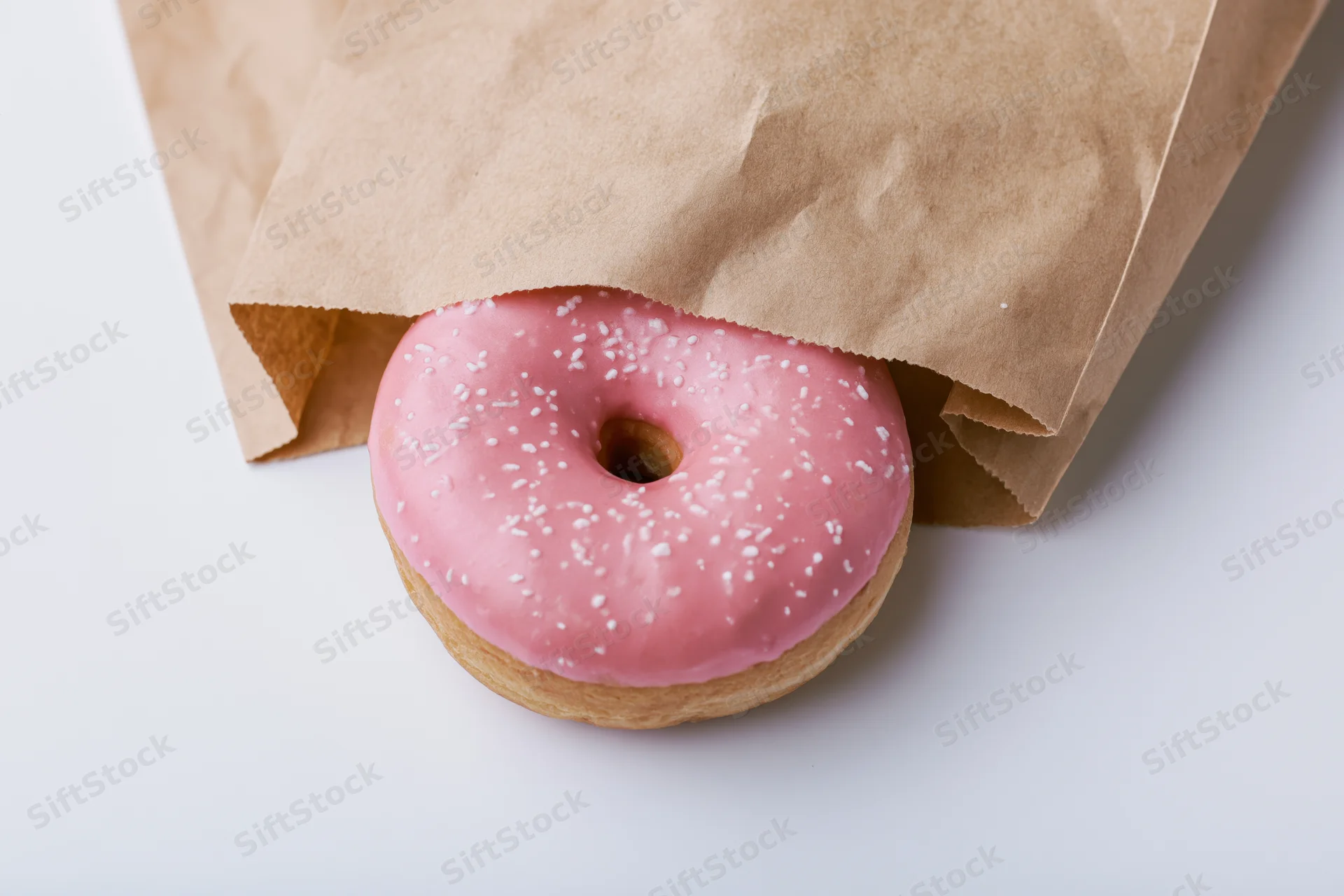 Pink glazed donut with white sprinkles peeking from a brown paper bag on white surface