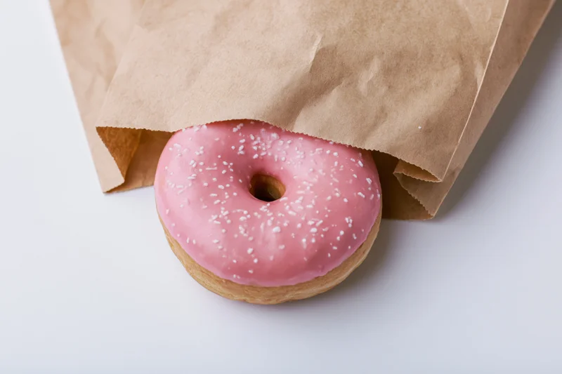 Pink glazed donut with white sprinkles peeking from a brown paper bag on white surface