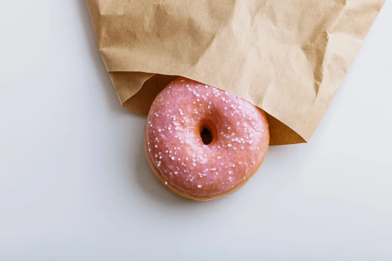 Pink glazed donut with white sprinkles peeking from a brown paper bag on white surface