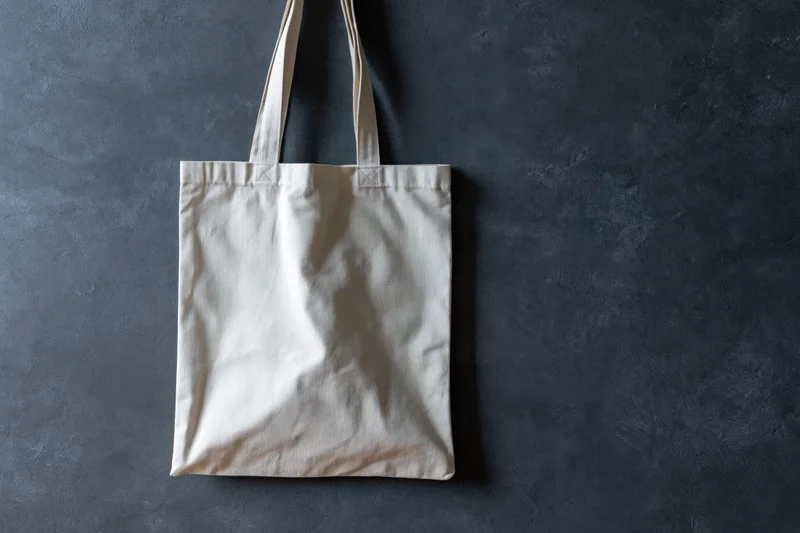 Plain white canvas tote bag hanging flat against dark textured wall