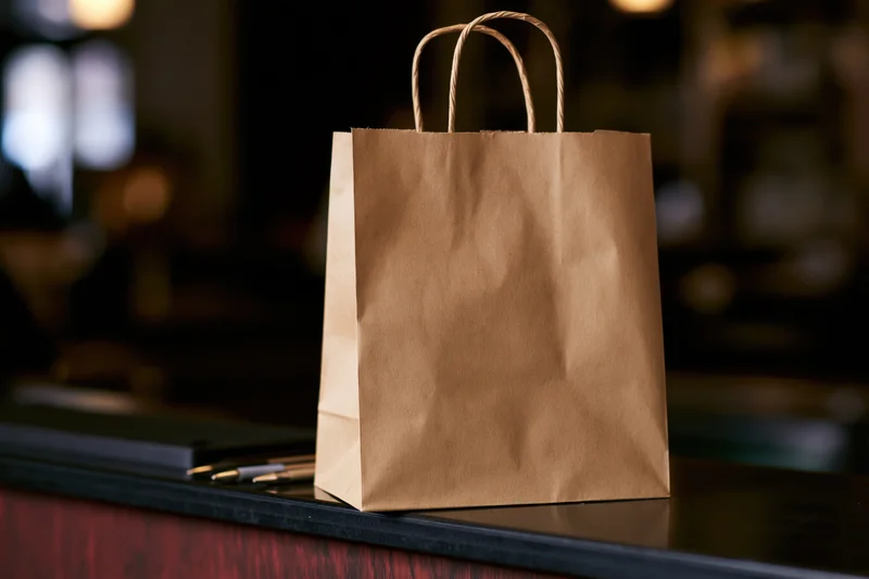 Brown kraft paper takeout bag with twisted handles sitting on a dark counter in a restaurant setting