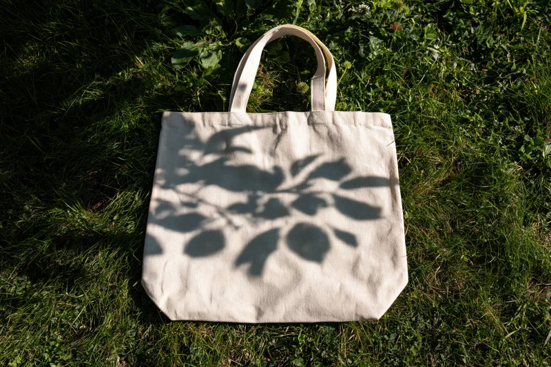 Cream canvas tote bag lying flat on green grass with leaf shadows from above