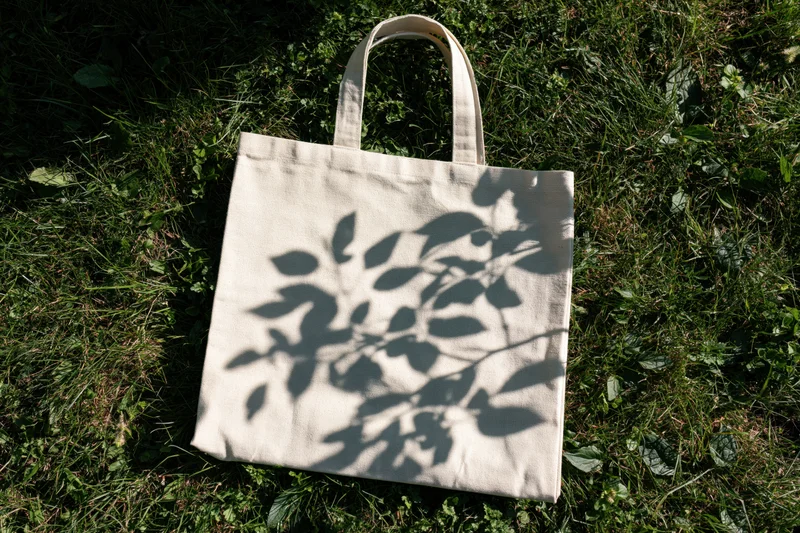 Cream canvas tote bag lying flat on green grass with leaf shadows overhead