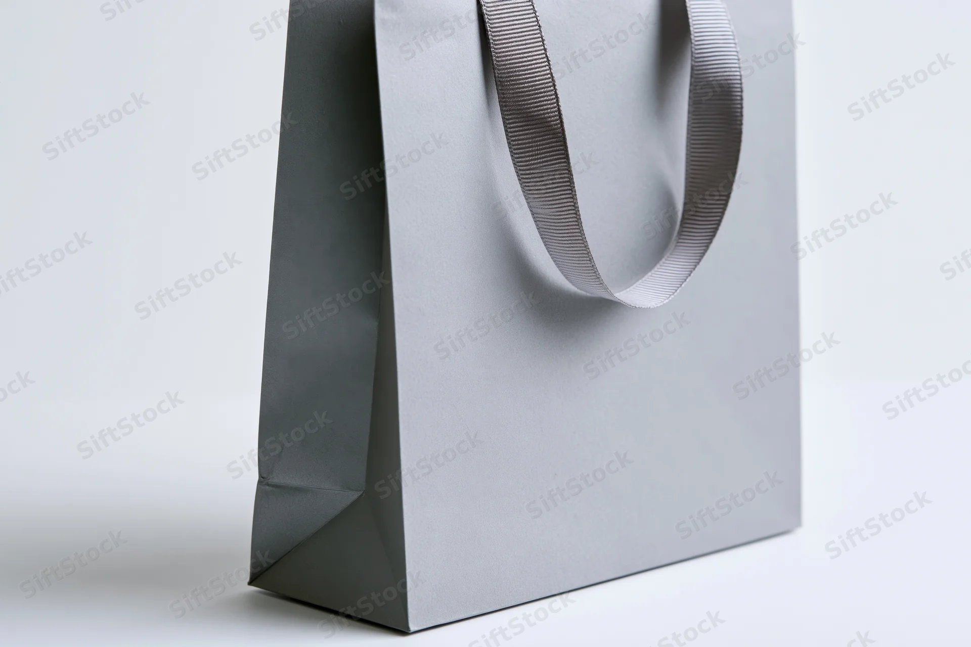 Grey paper shopping bag with ribbon handle standing upright on white surface