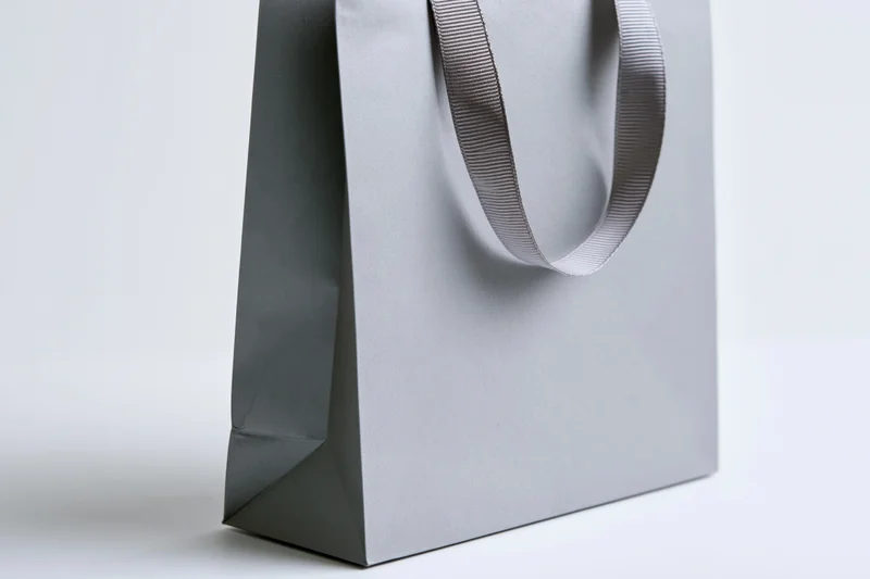 Grey paper shopping bag with ribbon handle standing upright on white surface