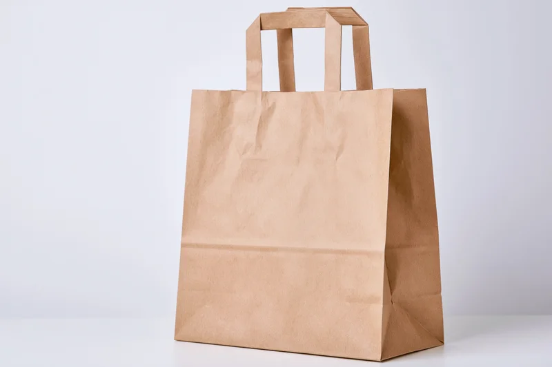 Plain kraft paper shopping bag with flat handles standing upright on white surface