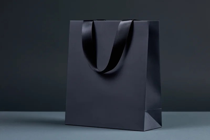 Matte black paper shopping bag with glossy ribbon handles on dark surface
