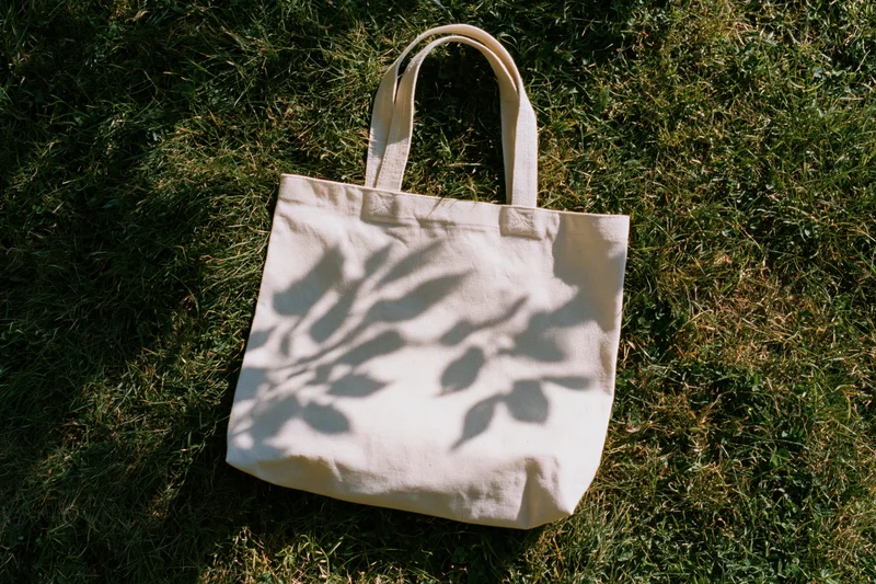 Off-white canvas tote bag lying flat on green grass with leaf shadows in sunlight