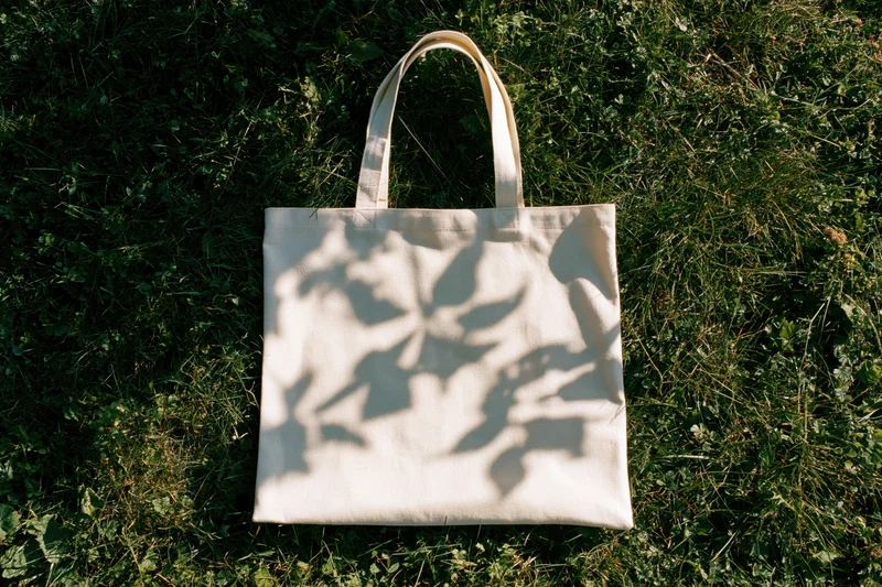 Off-white canvas tote bag lying flat on green grass with leaf shadows in sunlight