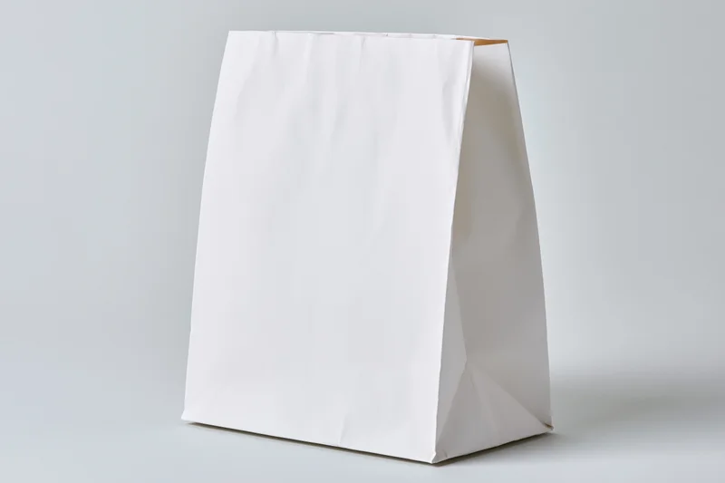 Plain white paper bag standing upright on a light surface with blank front for branding or design