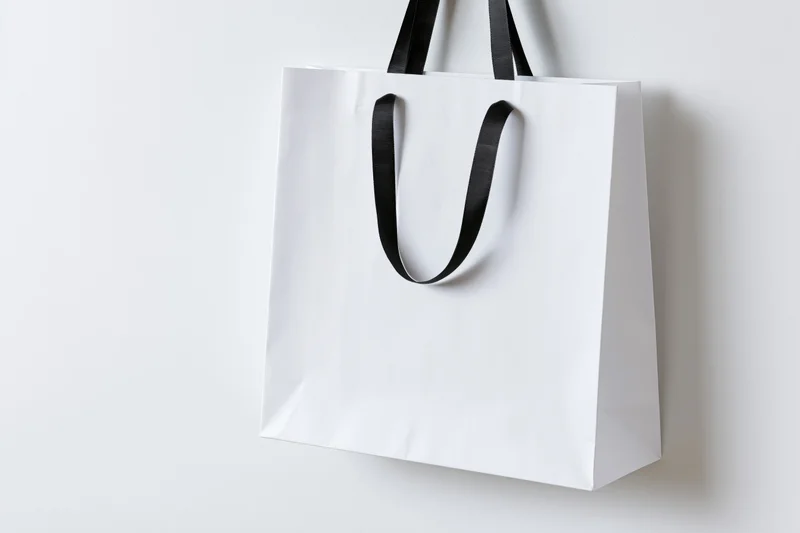 White paper shopping bag with black ribbon handles hanging against plain white background