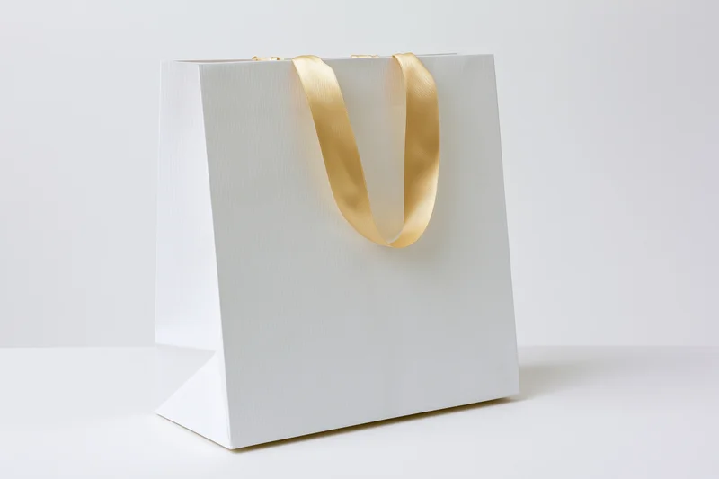 White paper shopping bag with gold satin ribbon handles on plain white surface