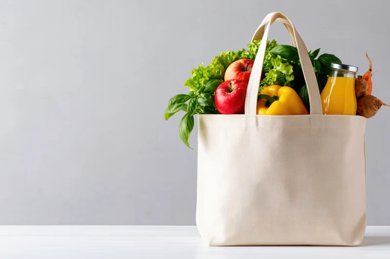 Reusable canvas tote bag filled with fresh fruits vegetables and juice on white surface