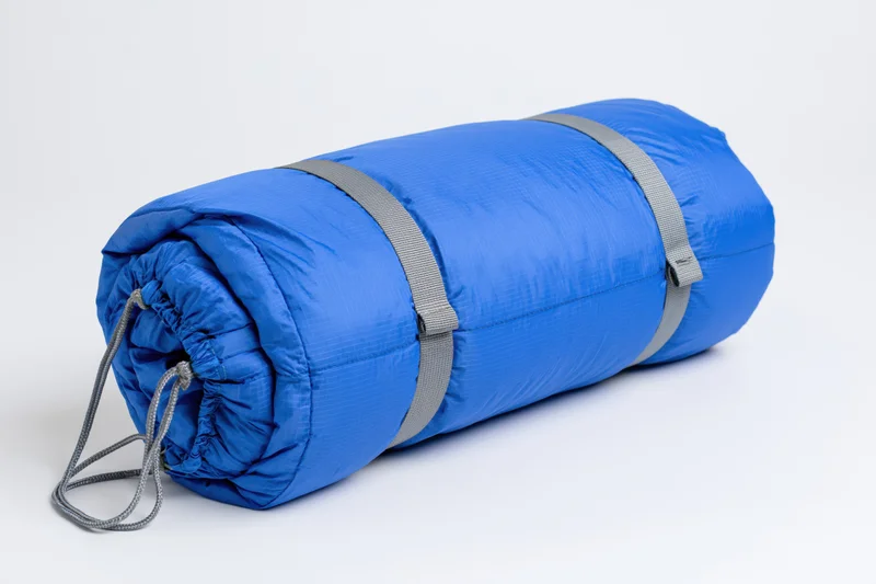 Blue nylon sleeping bag rolled tightly with straps and drawstring on white background
