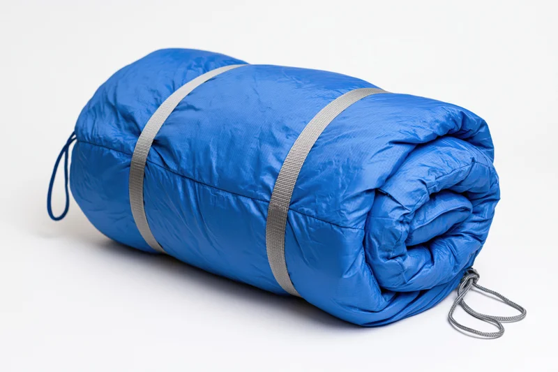 Blue nylon sleeping bag rolled tightly with straps on white background