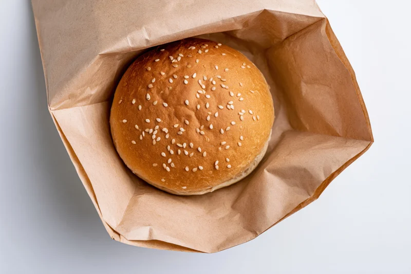 Sesame seed burger bun in open kraft paper bag on white surface