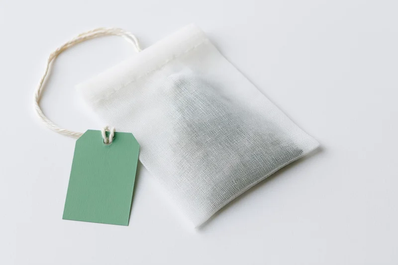 White mesh tea bag with green tag and string lying flat on white surface