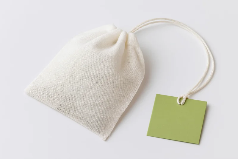 White mesh tea bag with green paper tag attached by string, lying flat on white surface