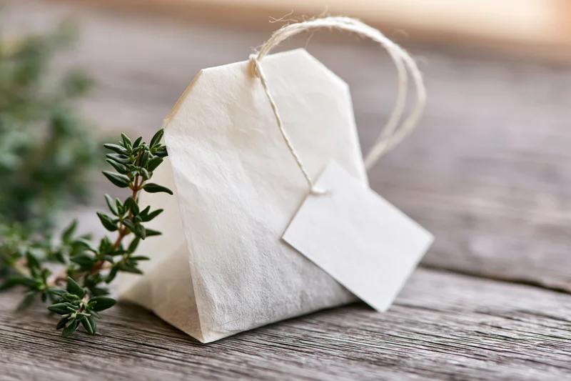 White paper tea bag with string tag and green herb sprig on wooden surface