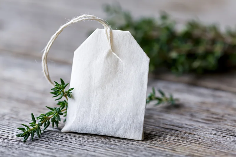 White paper tea bag with string tag resting on wooden surface beside fresh thyme sprigs
