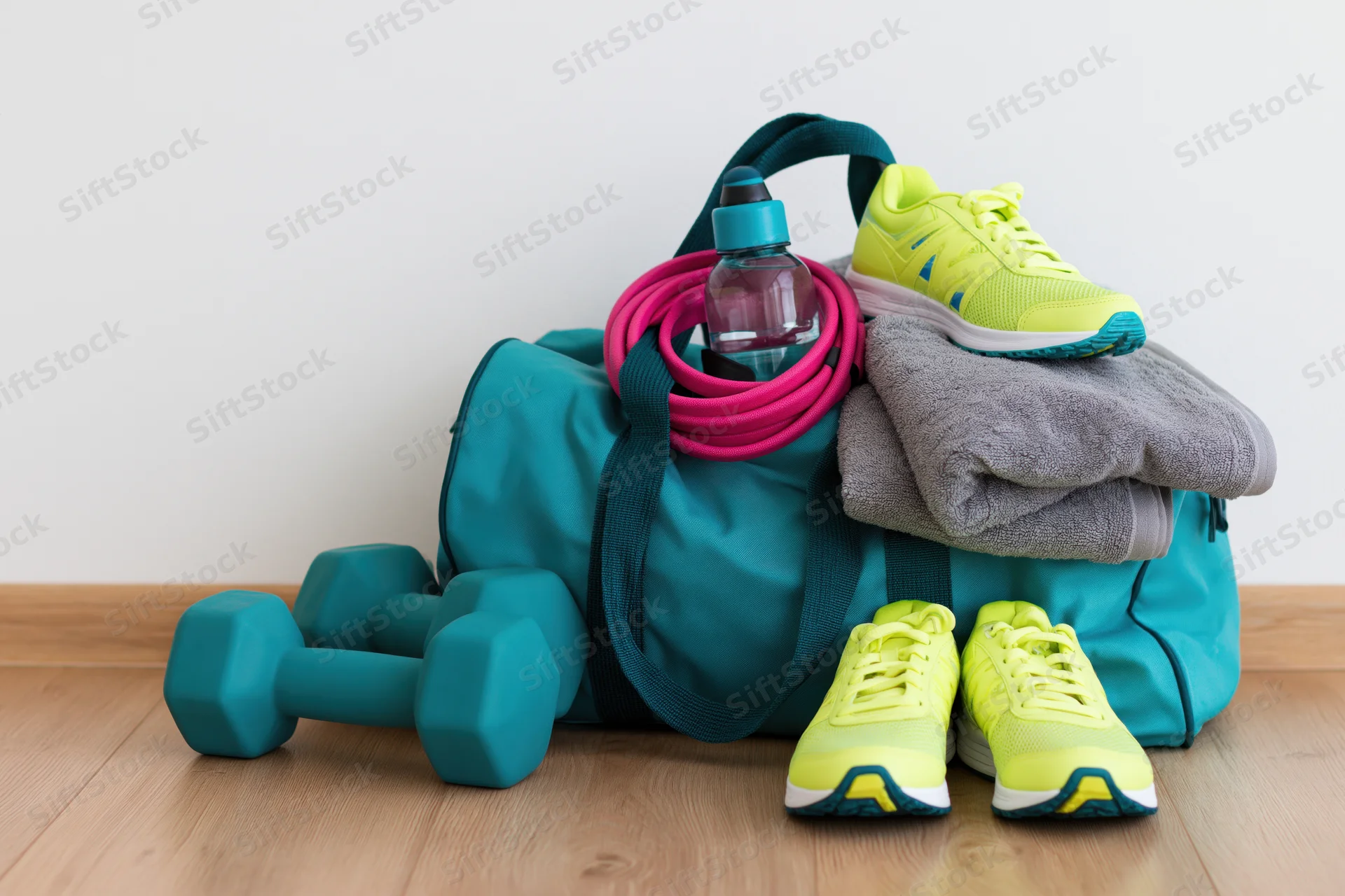 Teal gym bag overflowing with workout gear including neon yellow sneakers, dumbbells, resistance band and towel on wooden floor