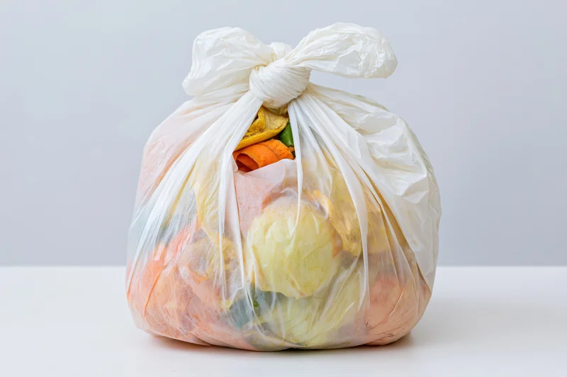 Tied white plastic bag filled with vegetable scraps and food waste on a white surface