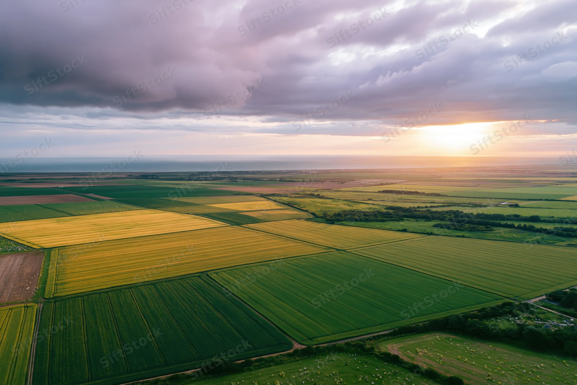 Aerial drone view of patchwork farmland fields under cloudy sky at sunset near ocean