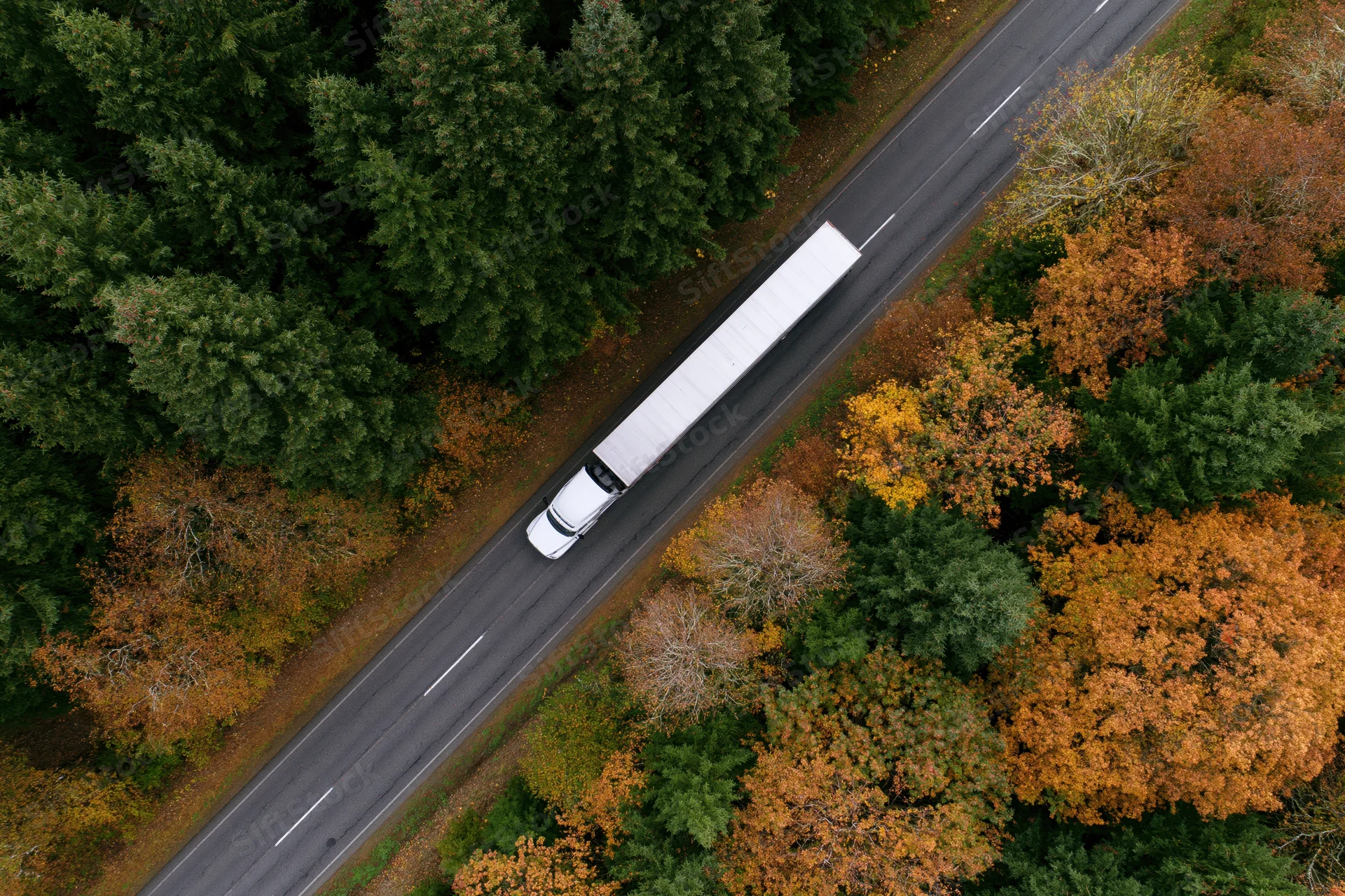 White semi-truck driving on a highway through an autumn forest from an aerial view