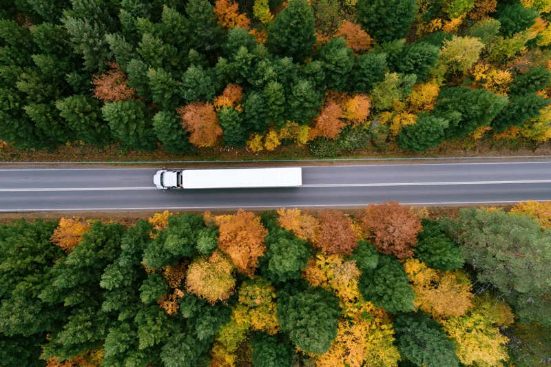 White semi-truck driving on highway through autumn forest from aerial view