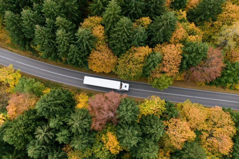 White semi-truck driving along a highway through autumn forest from aerial view