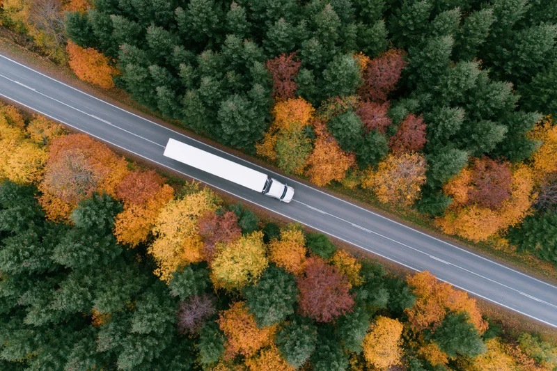 White semi-truck driving on highway through autumn forest from aerial view