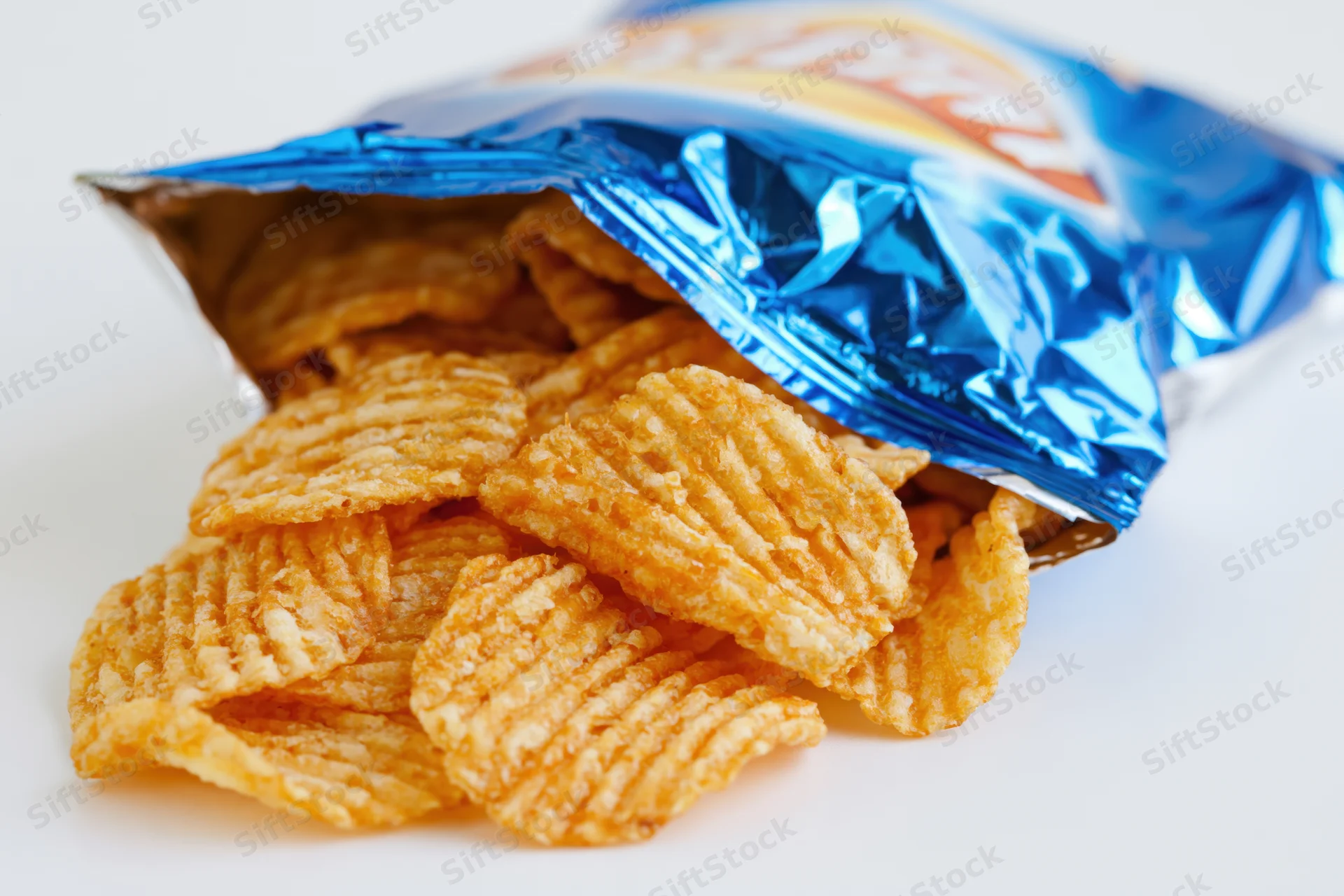 Ridged potato chips spilling from an open blue foil bag on white surface
