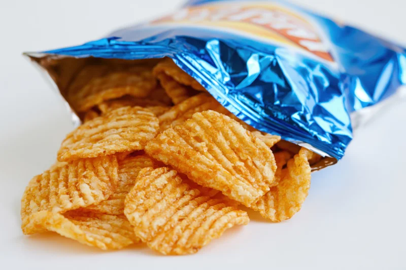 Ridged potato chips spilling from an open blue foil bag on white surface